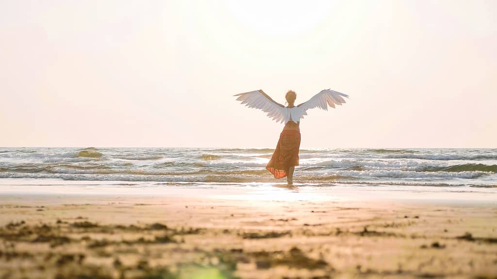Female winged angel on seashore in warm sunlight, rear view