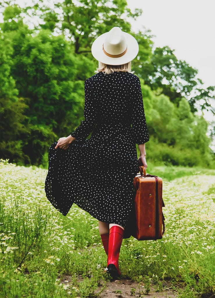 Blonde girl with suitcase and hat in chamomiles flowers field in summer time